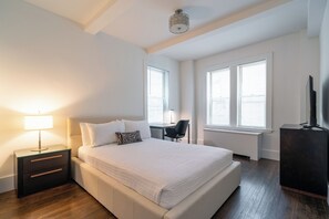 A bedroom with a bed, bedside table, lamp, desk, chair, and TV.