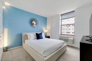A bedroom with a bed, a wall clock, a window, and a radiator. - Upscale Fully Furnished Two-Bedroom in Hoboken (Hoboken)