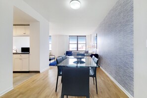 A modern dining area with a glass table and black chairs, a kitchen in the background, and a patterned wall.