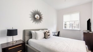Une chambre à coucher comprenant un lit, une table de chevet avec des tiroirs, une lampe, un miroir fixé au mur, une fenêtre et un téléviseur.
