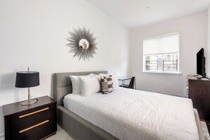 A bedroom with a bed, a nightstand with drawers, a lamp, a wall-mounted mirror, a window, and a TV.