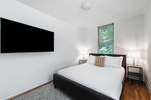 Une chambre moderne avec un grand lit, deux tables de chevet avec des lampes, un téléviseur à écran plat fixé au mur et une fenêtre avec des stores.