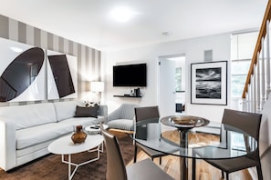 Un salon moderne avec un canapé blanc, une table à manger en verre et un téléviseur noir fixé au mur.