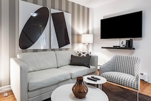 Un salon moderne avec un canapé gris, un fauteuil à motifs, une table basse agrémentée d’un vase et un téléviseur à écran plat fixé au mur.