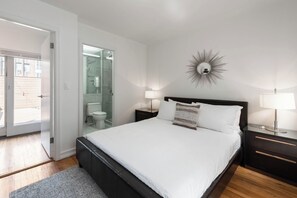 Un dormitorio con cama, mesitas de noche, un espejo y un baño visible a través de una puerta abierta.