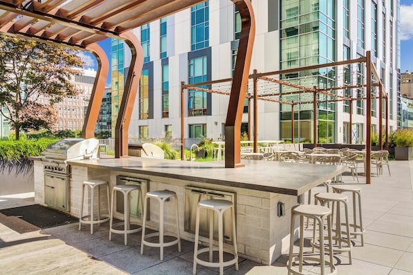 An outdoor bar area with a grill, bar stools, and a view of a modern building with large windows.