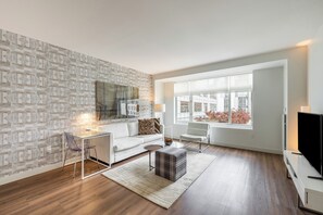 A modern living room with a white sofa, a glass coffee table, a plaid ottoman, and a flat-screen TV on a white stand.