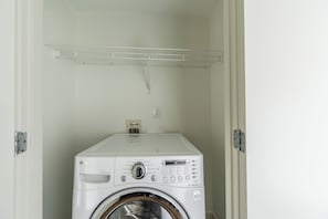 A compact laundry room with a front-loading washing machine, a metal shelf above, and a power outlet on the wall.