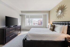 Quarto de hotel moderno com uma cama grande, televisão de tela plana, cômoda e vista da cidade por grandes janelas.
