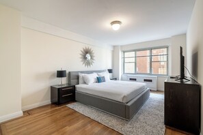 A modern bedroom with a large bed, a nightstand, a dresser, and a window with a view of buildings. - Elegant Murray Hill | Fitness Center & Laundry (New York)