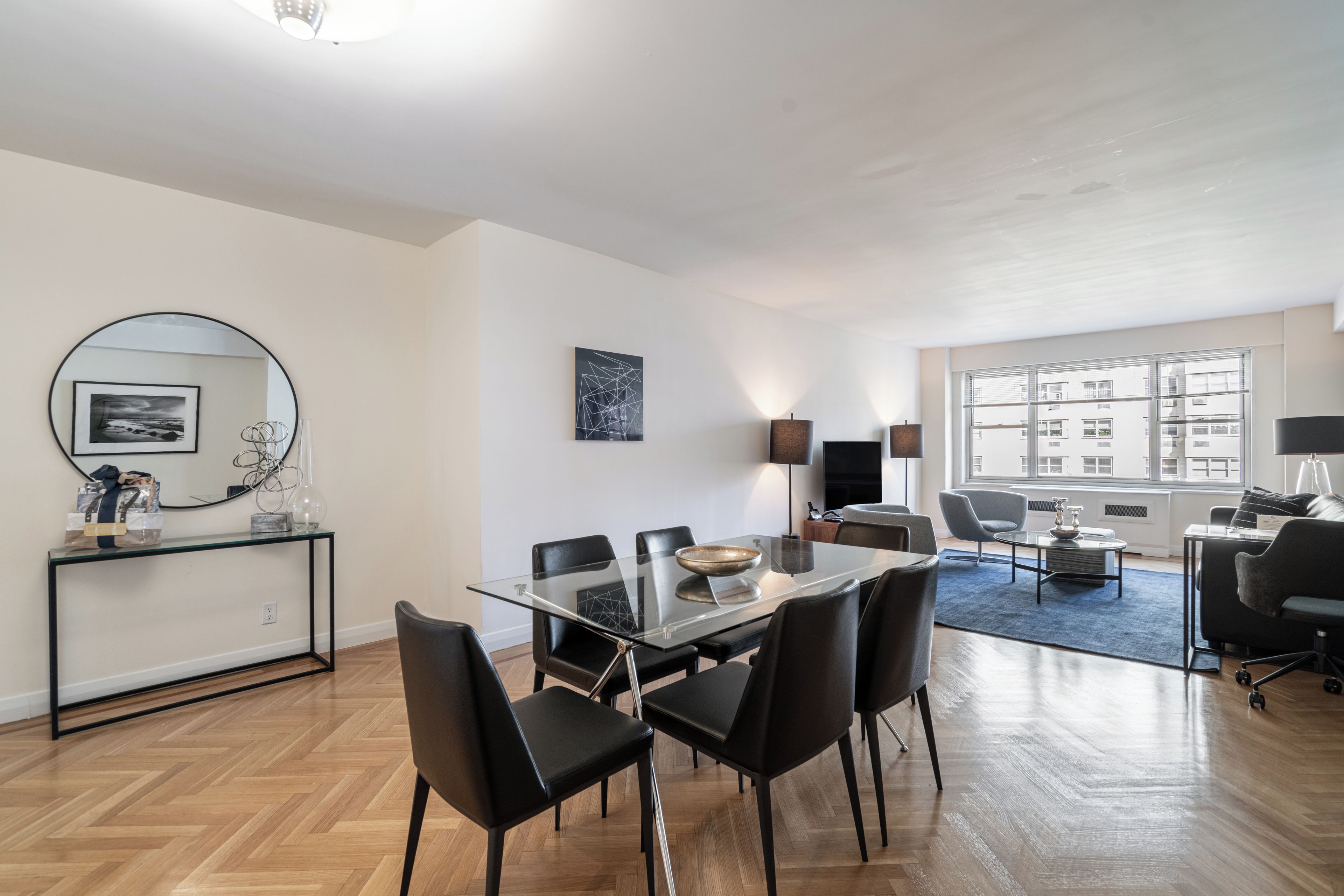 A modern living room with a glass dining table, black chairs, a blue rug, and a large window offering a city view.