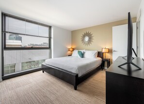 Une chambre d’hôtel moderne avec un grand lit, deux tables de chevet avec des lampes, un téléviseur posé sur un meuble, un miroir décoratif au mur et une vue sur la ville grâce à de grandes fenêtres.