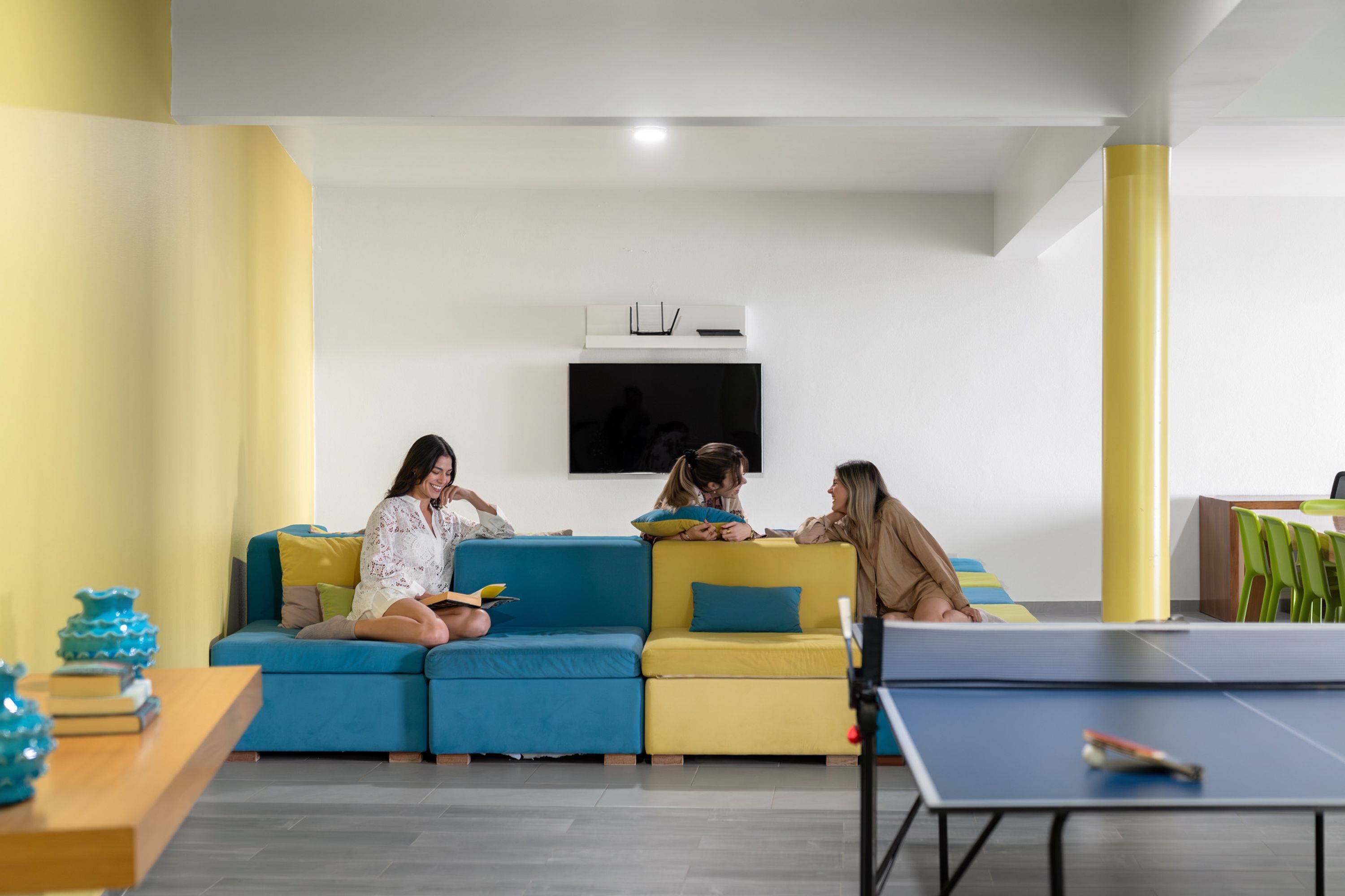 Una sala de estar moderna con un sofá azul y amarillo, un televisor de pantalla plana montado en la pared y una mesa de ping pong.