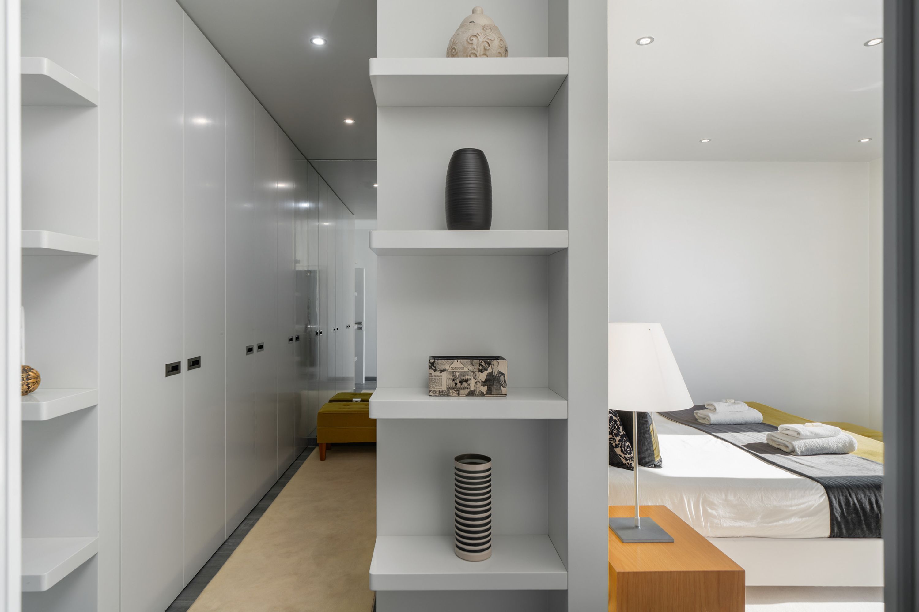 Une chambre d’hôtel moderne équipée d’un lit, d’une table de chevet, d’une lampe, ainsi que d’un couloir avec des casiers et des étagères.