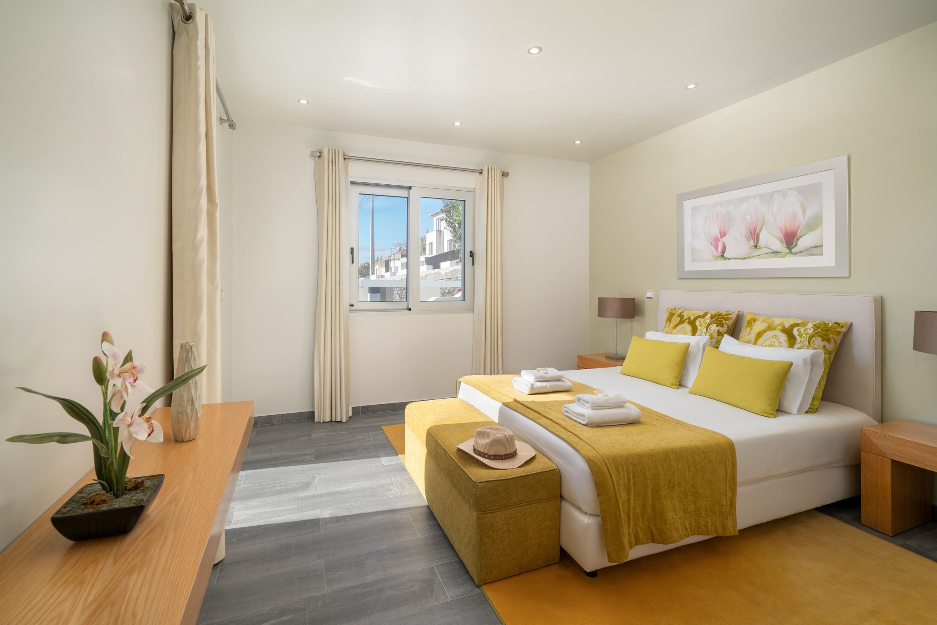 A modern hotel room with a bed, bedside tables, a window with curtains, a potted plant, and a painting on the wall.