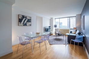 A modern living room with a glass dining table, clear chairs, a sofa, and a large window offering a city view.