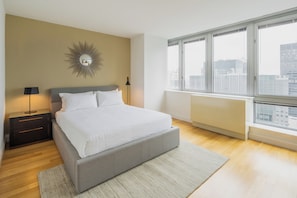 A bedroom with a bed, nightstand, lamp, and a large window with a city view.
