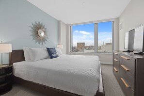 A modern hotel room with a large bed, a city view, a vanity with a mirror, and a flat-screen TV. - Gorgeous One Bed in Amenity-Rich Community (Boston)