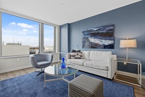 A modern living room with a glass coffee table, a white sofa, a blue rug, and a large ocean view painting on the wall. - Gorgeous One Bed in Amenity-Rich Community (Boston)