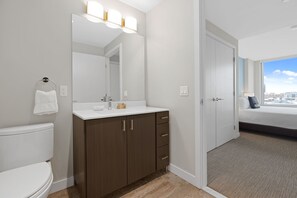 A modern bathroom with a white toilet, a vanity with a mirror, and a sink. There is a towel hanging on the wall and a door leading to a bedroom with a bed and a view of the city. - Gorgeous One Bed in Amenity-Rich Community (Boston)