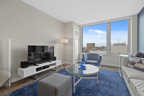A modern living room with a flat-screen TV, a glass coffee table, a blue vase, a grey armchair, and a sofa with a patterned throw.