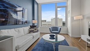 En moderne stue med sofa, sofabord i glas, en blå lænestol, fjernsyn og et stort vindue med udsigt over byen.