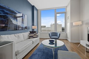 Un soggiorno moderno con un divano, un tavolino in vetro, una poltrona blu, una TV e un'ampia finestra con vista sulla città.