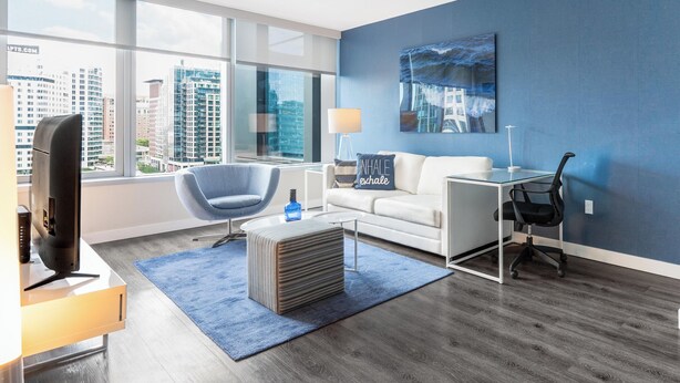 A modern living room with a white sofa, a blue armchair, a glass coffee table, and a blue rug. There is a painting on the wall and a lamp on a side table. - Spacious One Bedroom in Luxury Boston Building (Boston)