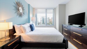 A modern hotel room with a bed, a TV, a nightstand, a lamp, and a view of the city through large windows. - Spacious One Bedroom in Luxury Boston Building (Boston)