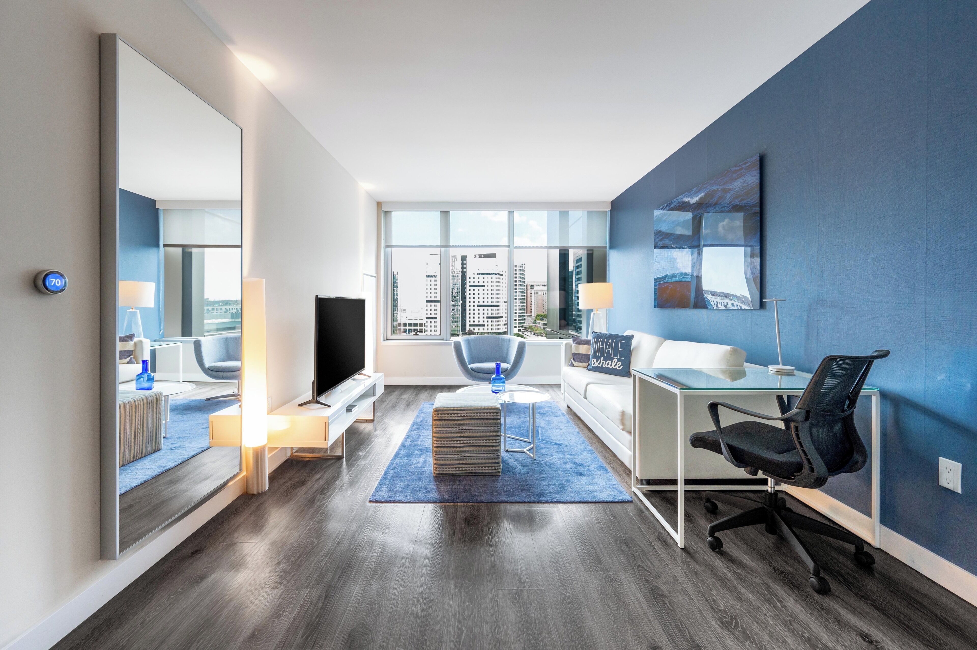 A modern hotel room with a large mirror, a flat-screen TV, a desk with a chair, a sofa, and a large window offering a city view.