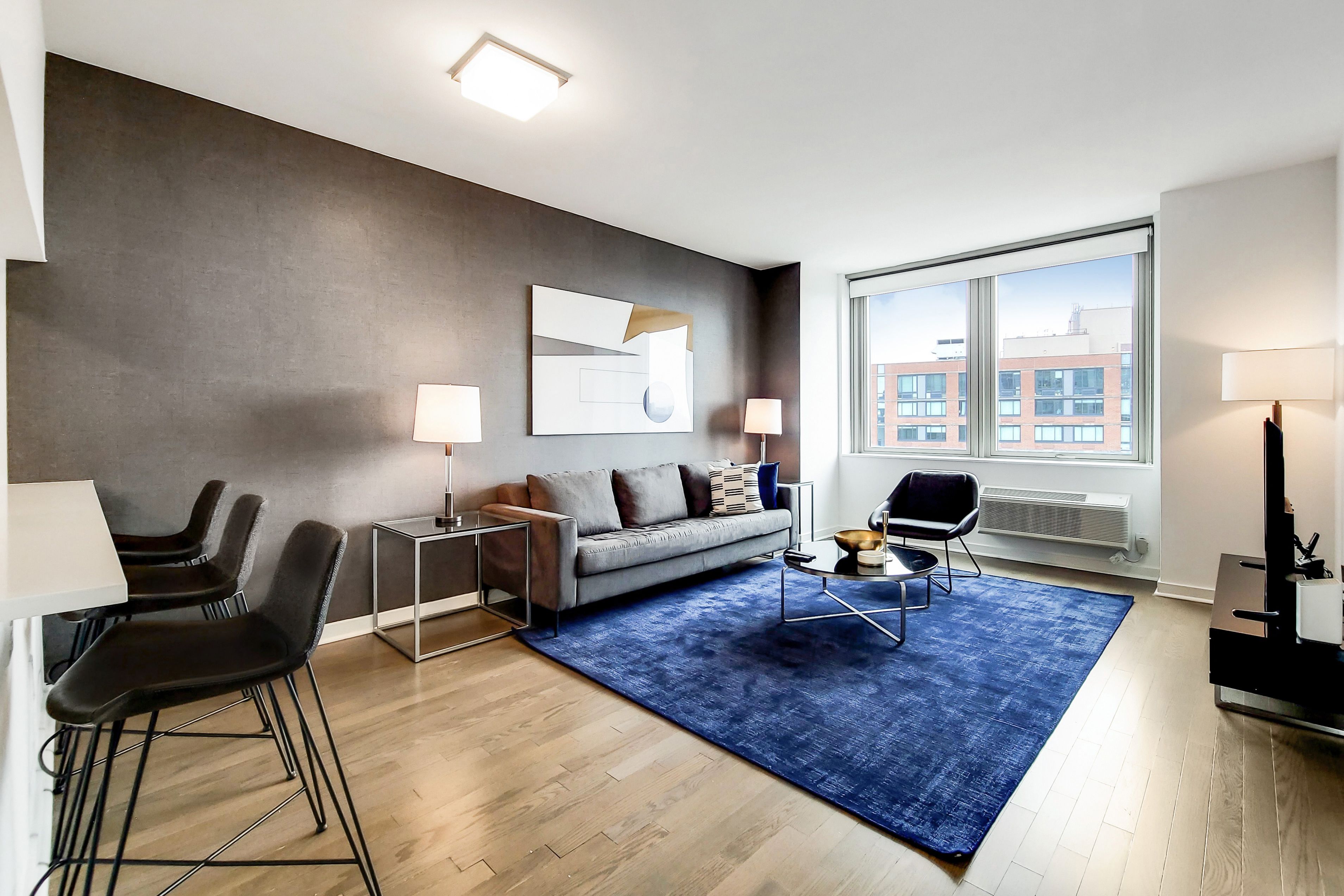 Un salon moderne avec un canapé, une table basse et des fauteuils, une grande fenêtre donnant sur la ville et un tapis bleu.