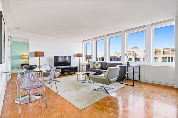 A modern living room with a flat-screen TV, glass dining table, and chairs, large windows with a city view, and a wooden floor.