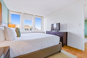 A bedroom with a large bed, a dresser, a television, and a window with a city view.
