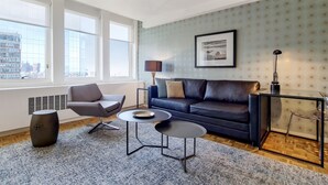Un salon moderne avec un canapé en cuir, deux tables rondes, une table d’appoint et une grande fenêtre donnant sur la ville.