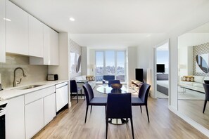 Une cuisine moderne avec des meubles blancs, un coin repas doté d’une table ronde en verre et de chaises noires, et une grande fenêtre offrant une vue sur la ville.