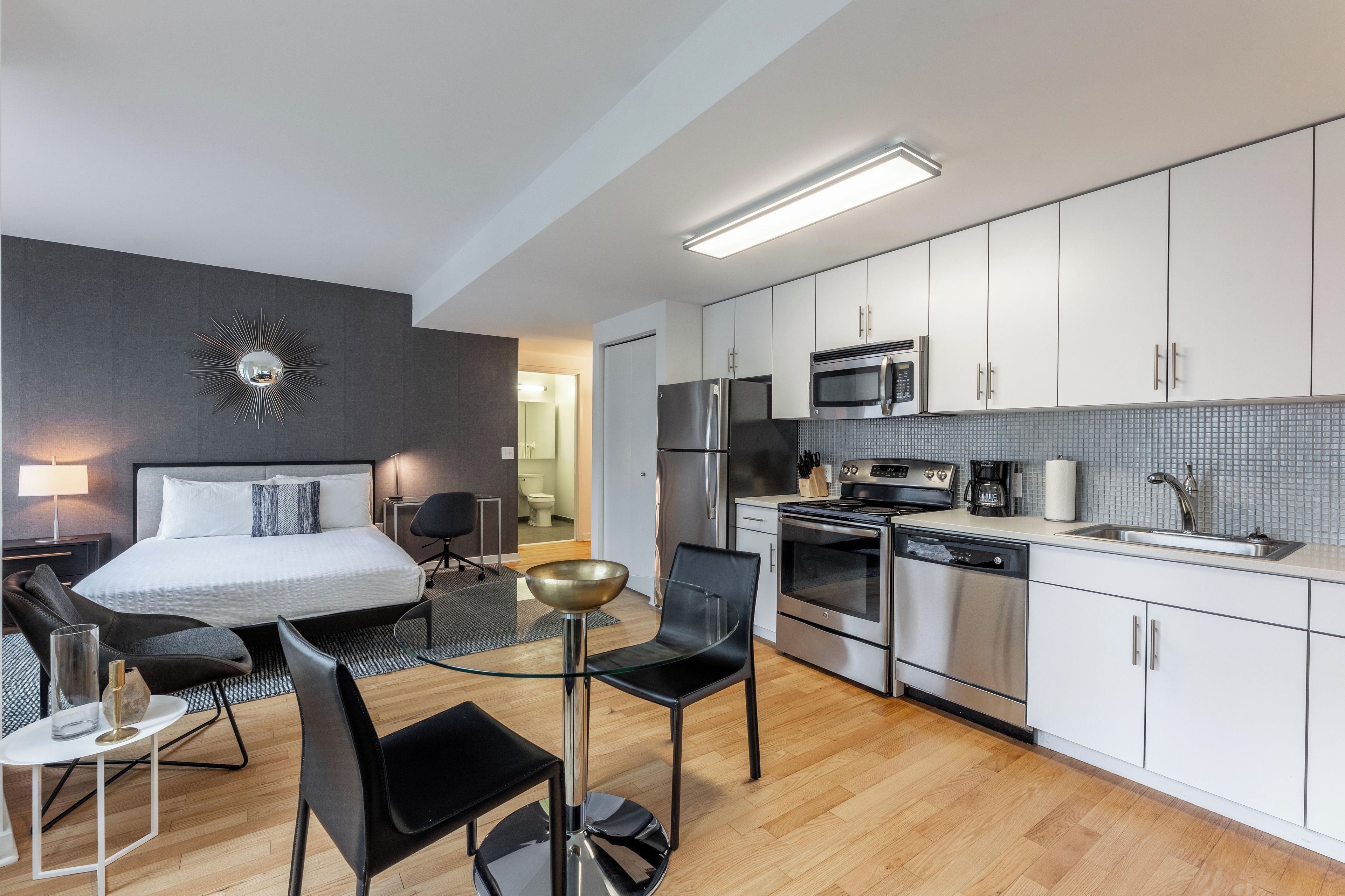 A modern hotel room with a bed, a dining area with a glass table, black chairs, a kitchen with white cabinets, a microwave, a refrigerator, and a dishwasher.