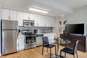 Een moderne keuken met roestvrijstalen apparatuur, een magnetron, een fornuis en een vaatwasser. Er is een eethoek met een glazen tafel en twee stoelen. Een flatscreen televisie is aan de muur bevestigd.