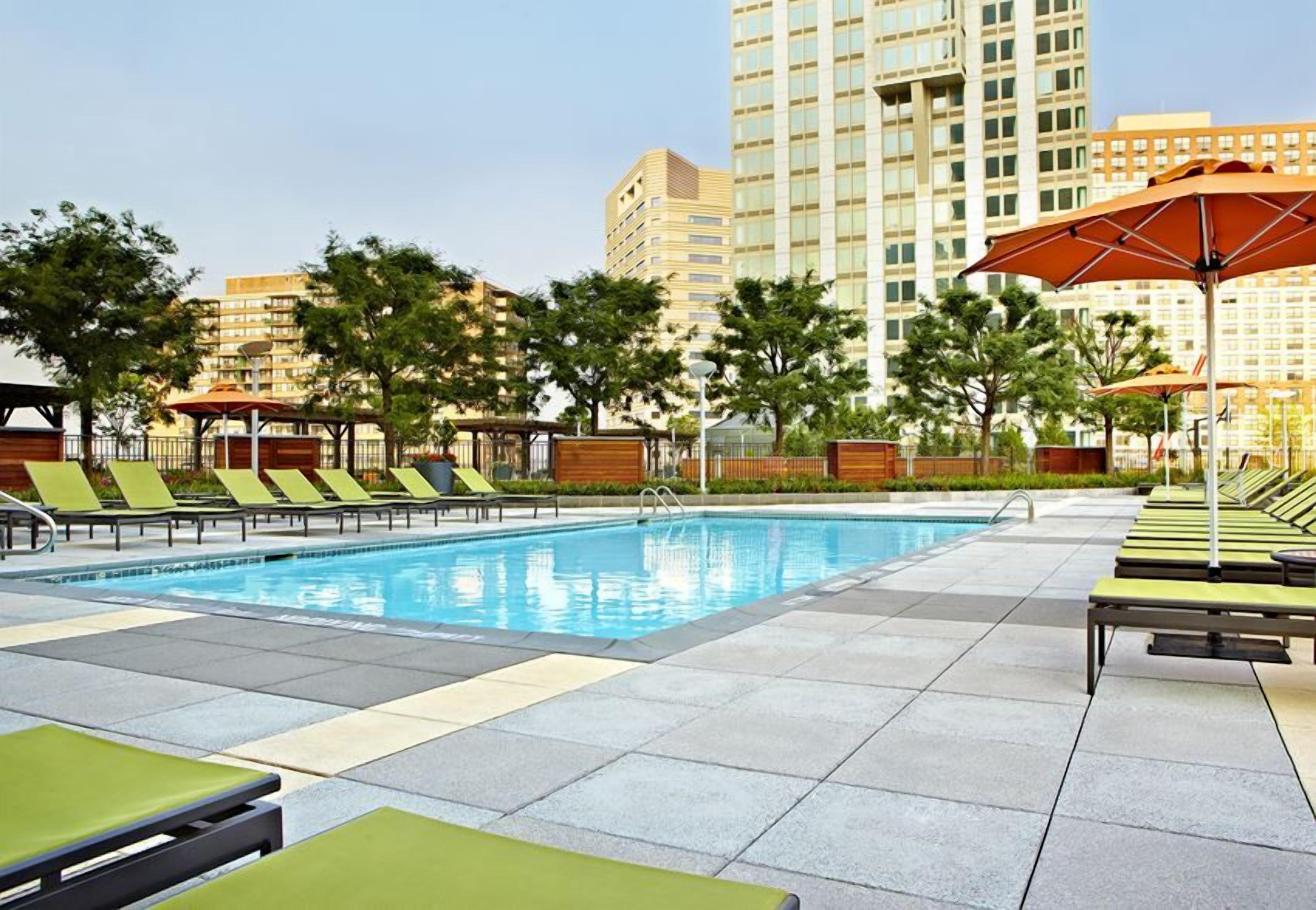 A swimming pool with lounge chairs and umbrellas, surrounded by trees and buildings.