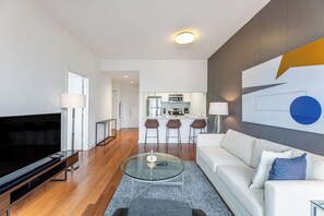 Un salon moderne avec un téléviseur à écran plat, une table basse en verre, un canapé blanc et une cuisine en arrière-plan.