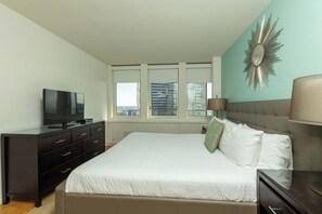 A bedroom with a large bed, a TV on a dresser, a window with a city view, and a decorative mirror on the wall. - One Bedroom Apartment in Back Bay Building (Boston)