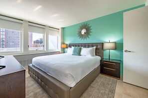A modern hotel room with a large bed, a city view, a sunburst mirror, and a nightstand with lamps. - One Bedroom Apartment W/ Luxury Amenities (Boston)