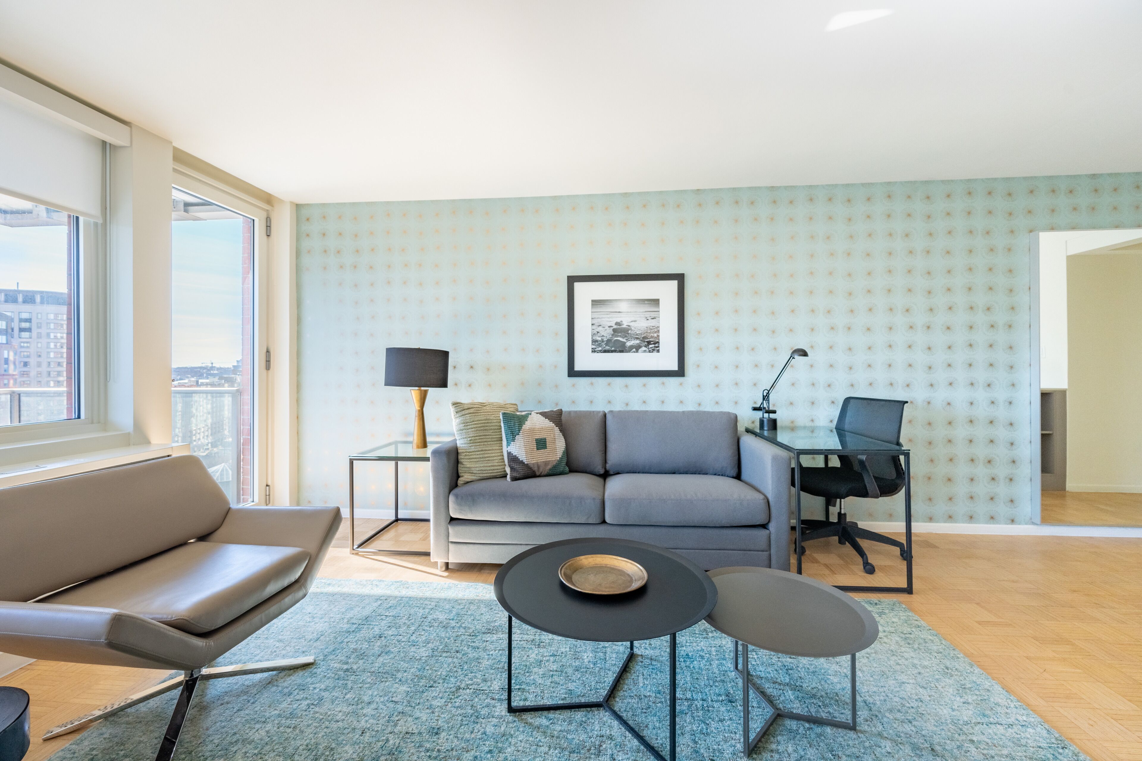 A modern living room with a grey sofa, a round black coffee table, and a black office chair. There is a framed picture on the wall and a lamp on a side table.