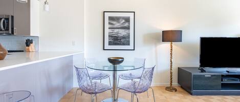 A modern kitchen with a microwave, a glass dining table with chairs, a framed picture on the wall, a floor lamp, and a TV on a cabinet.