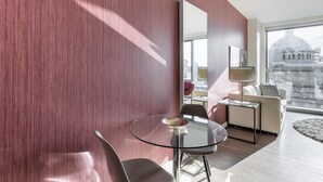 Ett modernt hotellrum med ett matbord i glas, två stolar, en soffa, en lampa och ett stort fönster med utsikt över staden.