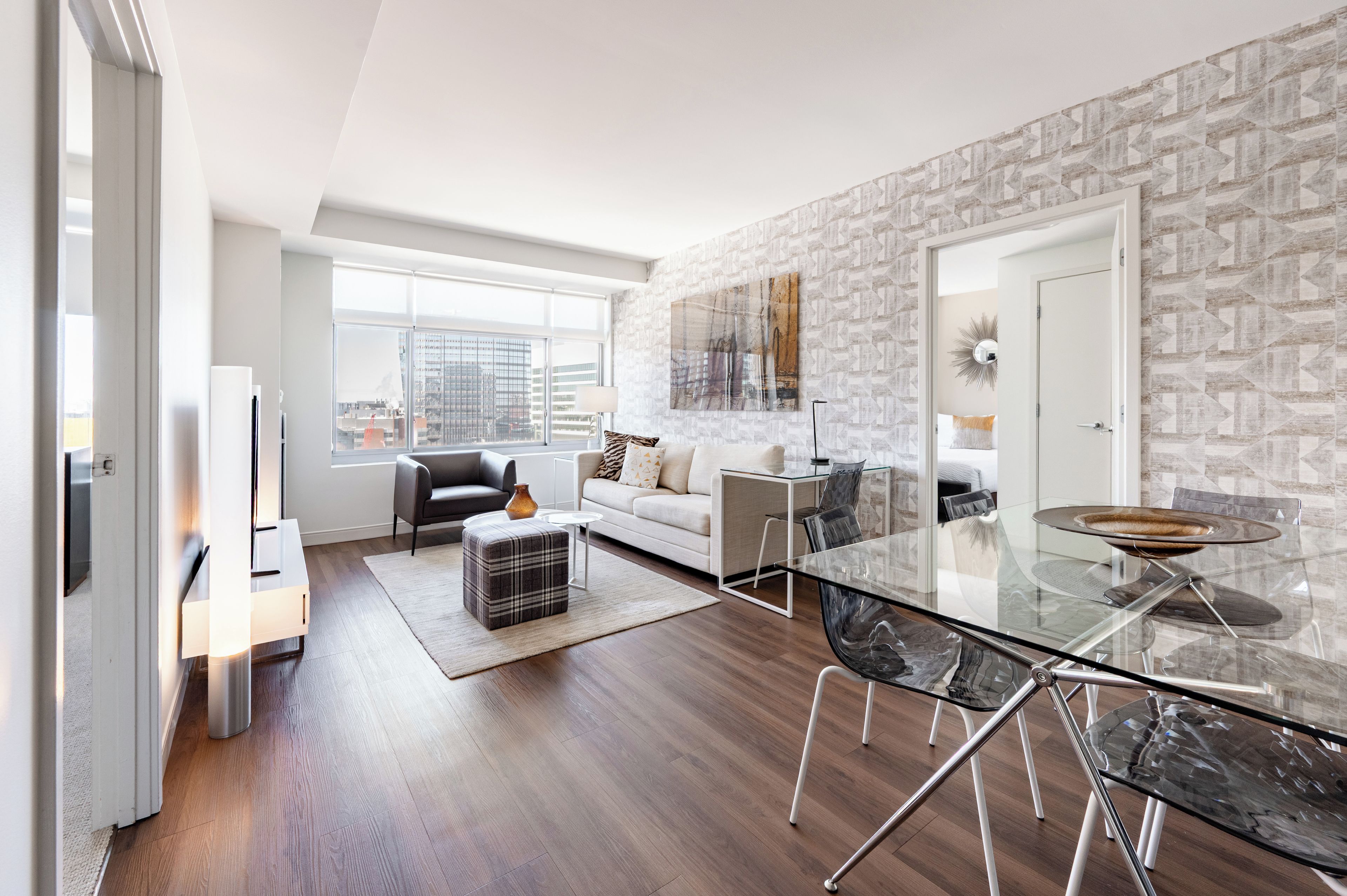 A modern living room with a glass dining table, a sofa, and a city view through large windows.