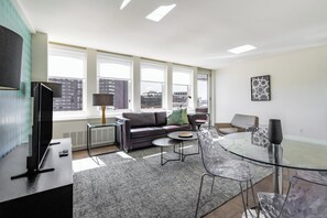 A modern living room with a sofa, coffee table, and glass dining table. Large windows offer a city view.