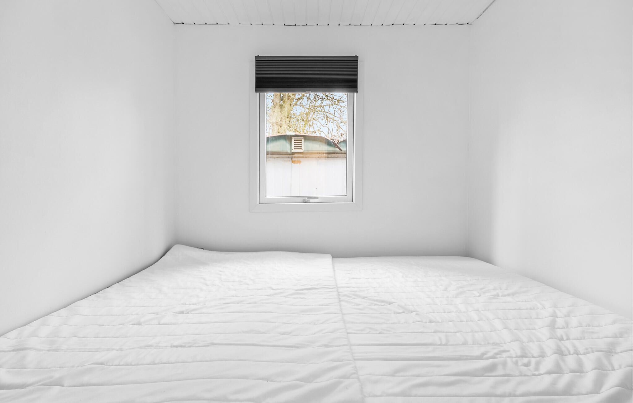 Een minimalistische slaapkamer met een enkel bed, een raam met zonwering en uitzicht op een boom en gebouw.