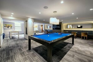 A pool table in a room with a carpeted floor, a television mounted on the wall, and a seating area with chairs.