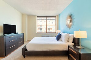 Un dormitorio con una cama, un aparador, un televisor, una ventana y un espejo montado en la pared.