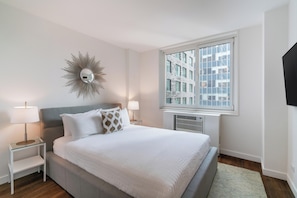 A modern bedroom with a large bed, a decorative mirror, bedside lamps, and a view of a building through a window.